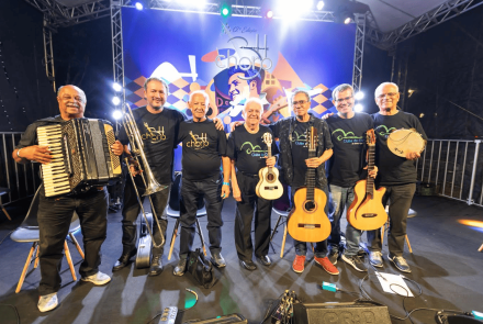 Fotografia colorida de sete músicos homens, adultos e idosos, posando sorridentes lado a lado no palco iluminado da 12ª Edição do BH Choro. Eles vestem calças e camisetas pretas do evento e seguram, da esquerda para a direita: um acordeon, um trombone de vara, um cavaquinho, dois violões e um pandeiro (o terceiro integrante está sem instrumento). Ao fundo, destaca-se um grande painel azul com a ilustração estilizada do músico Pixinguinha tocando flauta.