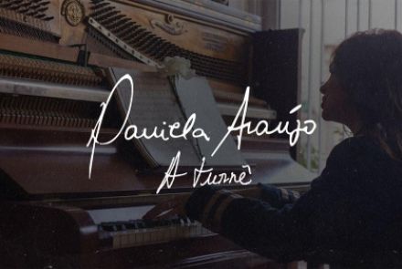 Cartaz horizontal em tons intimistas e rústicos. No lado direito, uma mulher de cabelos longos aparece de perfil, com os braços estendidos tocando as teclas de um piano vertical de madeira escura com o tampo superior aberto, revelando suas cordas e martelos internos. No centro, sobre uma partitura aberta adornada por uma flor branca em cima do piano, destaca-se o nome da artista "Daniela Araújo" escrito em uma caligrafia cursiva branca manuscrita, seguido pelo subtítulo "A Turnê" logo abaixo na mesma tipogr