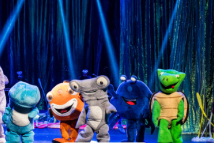 Foto de uma apresentação teatral infantil no palco. Ao centro, seis atores vestem fantasias grandes e coloridas de animais marinhos: um golfinho cinza, um peixe azul, um peixe-palhaço laranja, um tubarão-martelo cinza, um caranguejo azul-escuro e uma tartaruga verde. No canto direito, uma atriz caracterizada como pirata segura um instrumento musical. O cenário ao fundo tem cortinas brilhantes esverdeadas iluminadas por feixes de luz azul.