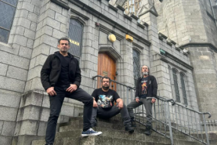 Fotografia colorida tirada de baixo para cima, mostrando três homens com roupas escuras e estilo casual/rocker posando nas escadarias de uma antiga construção de pedras cinzas, que lembra uma igreja gótica. Um homem está de pé à esquerda com a mão na cintura, outro está sentado ao centro dos degraus, e o terceiro está de pé à direita, perto de um corrimão de metal. Ao fundo, destacam-se uma porta de madeira em arco e janelas altas com vitrais.