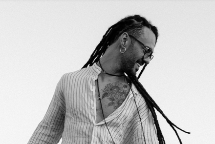 Fotografia em preto e branco com fundo claro. Um homem jovem, com pele negra clara, sorri de perfil olhando para baixo e para a direita. Ele tem cabelos longos com dreadlocks que caem pelas costas e ombros, usa óculos de grau, brinco de argola e um colar discreto. Veste uma camisa clara com listras verticais finas, que está entreaberta, revelando uma tatuagem de girassóis no peito. No canto inferior esquerdo, aparece uma pequena parte do braço de um violão.