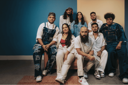 Foto em estúdio de um grupo de oito pessoas vestindo roupas jeans e brancas, posando juntas diante de uma parede dividida entre as cores azul e bege.