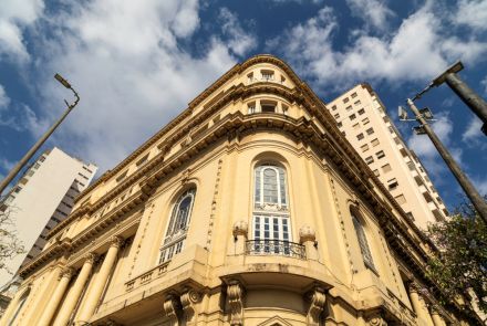 Edifício antigo de fachada bege com colunas e janelas altas em arco. O prédio ocupa a esquina, e ao fundo aparecem prédios residenciais modernos. Há postes de iluminação e parte de uma árvore no canto da imagem. O céu está azul com algumas nuvens brancas.