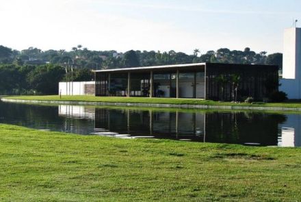 Edifício moderno e retangular no Parque Ecológico da Pampulha. O prédio, de cor escura, tem grandes janelas de vidro e uma torre branca ao lado. Ele está à beira de um lago que reflete sua imagem. O primeiro plano é uma vasta área de grama verde que se estende até a água.