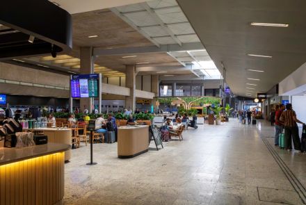 Corredor principal do aeroporto, com um café à esquerda onde passageiros estão sentados em mesas. O saguão tem piso de granito claro, teto de concreto aparente e pilares cilíndricos. Pessoas caminham com malas e aguardam em assentos distribuídos pelo ambiente.