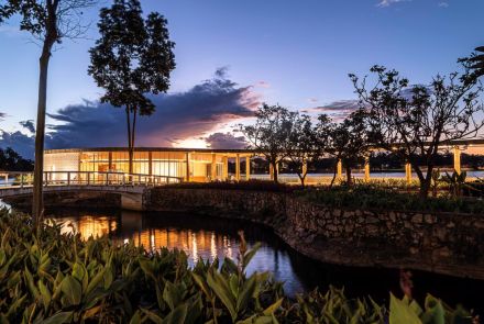 Casa do Baile ao anoitecer. É uma construção redonda e moderna, iluminada por dentro, que se estende sobre a água, conectada por uma ponte curva. O céu está em tons de azul, roxo e laranja do pôr do sol. A vegetação verde no primeiro plano reflete na água escura.