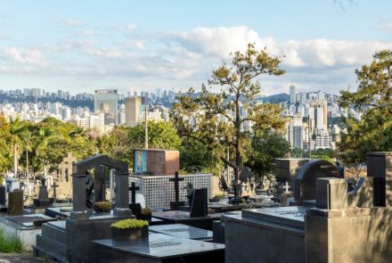 Um cemitério em uma tarde ensolarada. No primeiro plano, há vários túmulos de granito escuro, com alguns adornados por pequenas plantas verdes. As lápides têm diferentes formatos, algumas com esculturas e cruzes. No plano de fundo, erguem-se muitos edifícios e arranha-céus, formando um horizonte de cidade. Um edifício mais alto, de cor clara e com janelas espelhadas, destaca-se entre os demais. O céu, ao alto, é azul com algumas nuvens brancas, e a luz do sol ilumina a cena de forma clara.