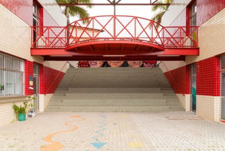 Entrada interna com escadaria larga de blocos cinzas que leva ao andar superior. Acima, há uma passarela vermelha metálica com grades. No fundo, parte de um mural colorido com rostos é visível.