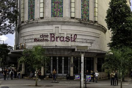 Edifício Cine Theatro Brasil, uma construção histórica em Belo Horizonte. No centro da foto, a fachada curva do prédio é visível. A entrada, ao nível da rua, possui grandes portas de vidro e metal. Acima da entrada, a fachada apresenta a inscrição "cine theatro Brasil" com letras de cor rosa, e detalhes arquitetônicos em relevo. Na parte superior da construção, grandes vitrais retangulares, com padrões coloridos em tons de verde, rosa e azul, se destacam. Em frente ao prédio, há palmeiras e pessoas.
