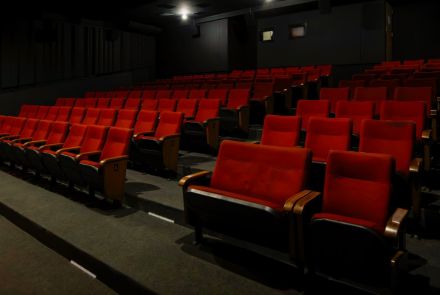 O interior de um cinema, com várias fileiras de poltronas vazias e vermelhas. O ambiente é escuro, iluminado apenas por algumas lâmpadas no teto. As poltronas são de veludo vermelho e têm apoios de braço de madeira escura.