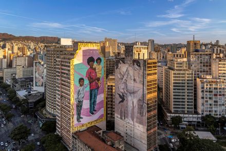 Vista aérea do centro de Belo Horizonte, destacando dois murais em prédios vizinhos: à esquerda, uma mulher negra com duas crianças em cores vibrantes; à direita, duas figuras femininas nuas sobrepostas, sendo que a da frente usa máscara e segura uma ave. A cena é emoldurada por arranha-céus e pela Serra do Curral ao fundo.