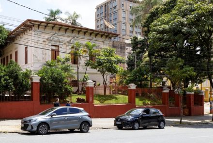 Imagem mostra a fachada de um casarão antigo de dois andares, pintado de branco com detalhes em vermelho, cercado por muro e grades também vermelhas. Em frente ao prédio há árvores, arbustos e duas pessoas caminhando na calçada. Dois carros estão estacionados na rua, um cinza e um preto. Ao fundo aparece um prédio moderno de vários andares e outras árvores grandes. Fios elétricos atravessam a parte superior da cena.
