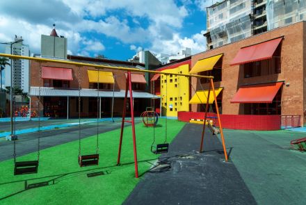 Pátio externo com um balanço de estrutura vermelha e laranja em primeiro plano, sobre uma área de grama sintética. Ao fundo, a fachada da escola em tijolo aparente, com toldos vermelhos e amarelos, e um bloco central amarelo com pequenas janelas quadradas. Prédios altos cercam o local.