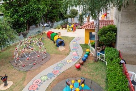 Vista de cima de um grande parquinho gramado com diversos brinquedos. Um caminho de cimento com letras coloridas do alfabeto serpenteia pelo jardim. Há um túnel em formato de centopeia, um gira-gira, um domo de escalada e uma casinha de brinquedo amarela com telhado de cerâmica.