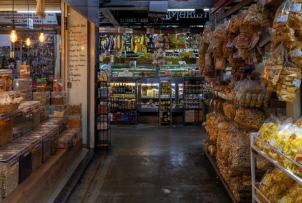 Interior de uma loja ou mercado, cheia de produtos. O corredor principal é estreito, com bancas e prateleiras dos dois lados. À esquerda, há uma variedade de cereais, grãos e nozes em recipientes de vidro transparente. À direita, prateleiras estão lotadas de pacotes plásticos transparentes com salgadinhos, biscoitos, pães de queijo e doces.