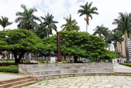 Em primeiro plano, um palco com três degraus feitos de cimento, com o chão à frente coberto por lajes de pedra irregular. Acima, no centro, uma escultura escura, de formato orgânico e retorcido, emerge entre duas árvores de copa densa e verde. Ao fundo, muitas palmeiras altas e edifícios