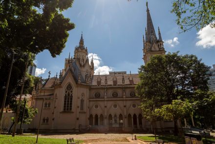 A Igreja de Nossa Senhora da Boa Viagem, uma construção alta e imponente em estilo gótico. O edifício é de cor bege e possui uma fachada rica em detalhes arquitetônicos. No topo, há duas torres altas e pontiagudas. A fachada possui um grande vitral e várias janelas redondas. Em frente à igreja, há um gramado verde e um caminho de tijolos.