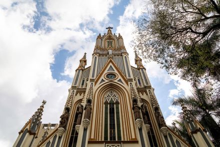 A vista de baixo para cima da fachada da Basílica Nossa Senhora de Lourdes. A igreja tem uma arquitetura gótica e é feita de pedra clara, com listras verticais em um tom mais escuro. A fachada é ricamente detalhada, com esculturas de santos em nichos, uma grande porta de entrada em arco e janelas longas. No topo da igreja, há uma torre pontiaguda com um relógio redondo no centro e uma cruz no topo. 