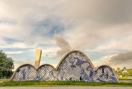 Uma igreja com estrutura arquitetônica de forma ondulada, coberta por azulejos azuis e brancos em padrão de mosaico, representando cenas históricas e figuras humanas. Ao fundo, um céu nublado com tons de cinza e azul claro. À esquerda, uma torre retangular de concreto. Em primeiro plano, grama verde e uma calçada. Palmeiras e árvores ao longe.