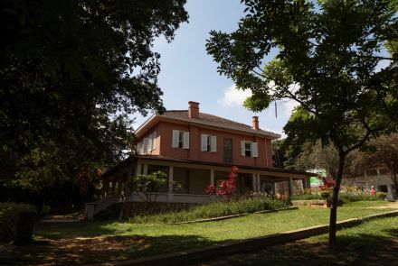 Imagem de uma casa de dois andares pintada de vermelho com janelas brancas. O telhado é de telhas marrons e há varanda no térreo. O jardim ao redor tem grama, plantas e flores coloridas. Árvores grandes envolvem a casa.