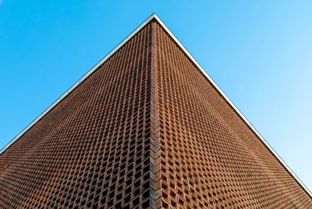 Vista de baixo de um canto do Mercado Novo em BH. A fachada tem uma textura de brises ou cobogós marrom-avermelhados, formando uma grade de losangos abertos que sobem em duas paredes. O céu azul claro emoldura a parte superior.