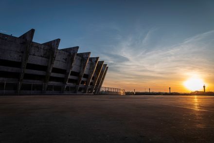 Imagem de um estádio de concreto visto ao entardecer, com destaque para sua estrutura monumental em forma de colunas inclinadas na fachada. O sol se põe no horizonte à direita, iluminando o céu em tons de laranja, dourado e azul, refletindo suavemente no piso amplo e vazio em frente ao estádio.