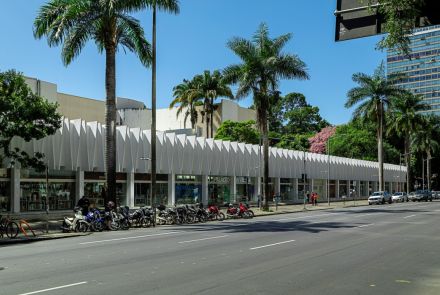 Fachada frontal do Palácio das Artes. O prédio tem uma estrutura longa e horizontal, com grandes janelas de vidro e uma parede branca em ziguezague no andar de cima. Uma rua larga está em primeiro plano, com motos e carros estacionados na calçada, que é ladeada por palmeiras. O céu está azul e sem nuvens.