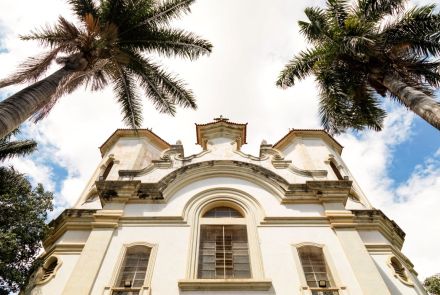 de baixo pra cima da Paróquia Santa Teresa e Santa Teresinha. A igreja, de cor clara, tem um estilo clássico com janelas em arco. No primeiro plano, duas grandes palmeiras com troncos grossos enquadram a igreja. O céu está azul com grandes nuvens brancas e claras.