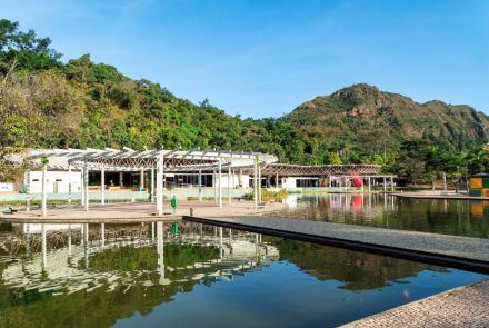 Área aberta com estruturas brancas em pergolado junto a um lago que reflete a paisagem. Ao fundo, construções baixas e vegetação densa, com uma montanha coberta por árvores e trechos de rocha, sob céu azul claro.