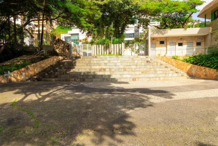 : Escadaria larga de pedra ocupa o centro da imagem, composta por degraus retos e lisos. Nas laterais, muros inclinados de pedra acompanham a subida. No alto, há um portão metálico fechado, atrás do qual se vê vegetação e parte de um prédio. À direita, um prédio baixo com portas e janelas dá vista para o pátio.