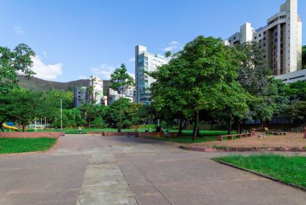 Vista ampla do parque, mostrando um pátio de cimento em primeiro plano, ladeado por áreas gramadas com bancos e árvores. Algumas pessoas estão sentadas nos bancos e uma bicicleta está estacionada. Ao fundo, o cenário é composto por prédios altos e uma serra coberta por vegetação.