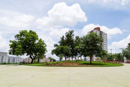 Grande área pavimentada de concreto clara, rodeada por árvores verdes. No centro há um gramado circular com canteiros de flores vermelhas. Ao fundo, edifício alto residencial.