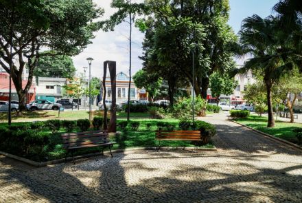 Uma praça com piso de pedras portuguesas e canteiros de grama e arbustos. Dois bancos de madeira com pés de ferro estão posicionados de frente, um à esquerda e outro à direita. Ao centro, entre árvores e postes de iluminação, há uma escultura de metal vertical e enferrujado. Ao fundo, veem-se ruas e casas.