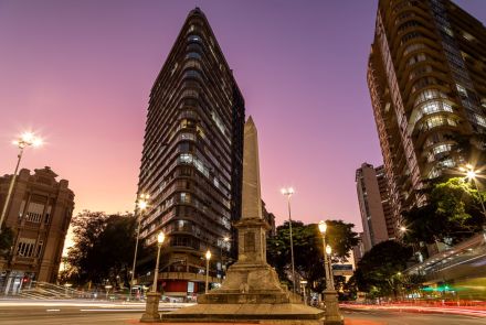 O obelisco da praça está centralizado, visto de baixo para cima contra um céu em dégradé de laranja, rosa e roxo durante o entardecer. Ao fundo, altos edifícios o cercam. Na rua, rastros de luz dos faróis dos carros em movimento indicam o trânsito intenso no cruzamento das avenidas.