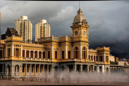 O histórico edifício da estação, de cor amarela, é visto em um ângulo próximo. A torre do relógio e sua cúpula se destacam contra nuvens escuras. Em primeiro plano, jatos de água de uma fonte no chão criam uma névoa branca que cobre a base do prédio, dando um efeito etéreo à cena.