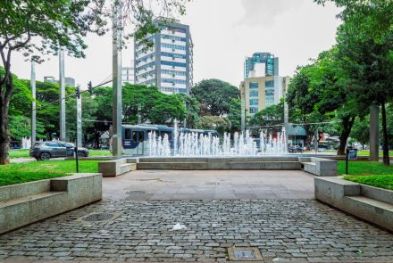 Praça com chafariz central, jatos de água brancos saem de um tanque retangular cercado por bancos de concreto. Ao fundo, prédios altos, ônibus e árvores verdes. Piso de pedra e concreto ocupa o primeiro plano.