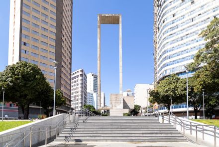 Vista a partir da base de uma longa escadaria de granito que leva à praça. O grande monumento de concreto em formato de portal está centralizado no topo da escada, enquadrado por dois altos edifícios nas laterais. O céu está limpo e com uma tonalidade de azul claro e uniforme.