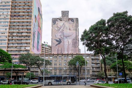 Vista frontal da praça, com canteiro de plantas verdes e árvores altas. Ao fundo, grande mural em prédio mostrando figuras humanas. À esquerda, prédio com fachada clara e outro com mural colorido. Ônibus e carros passam na avenida em frente.
