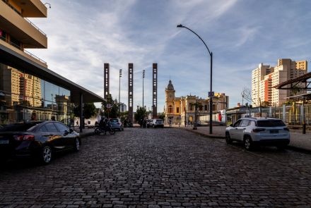 Vista de rua larga, com paralelepípedos e carros estacionados dos dois lados. À esquerda, um prédio histórico de tom amarelo, com sacadas e janelas trabalhadas. Ao fundo, aparecem três torres modernas mais altas, criando contraste entre o antigo e o novo.