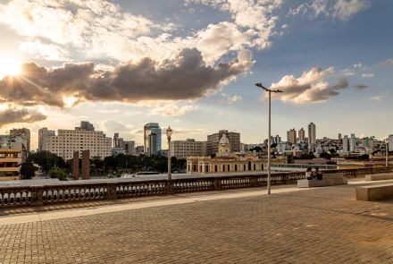Vista panorâmica da Praça da Estação, no muro da Rua Sapucaí. Em primeiro plano, o mirante da Rua Sapucaí com seu piso de blocos de pedra e um gradil de cimento. O sol poente à esquerda cria um brilho intenso e ilumina as nuvens. Ao fundo, o edifício da Estação e o horizonte de prédios.