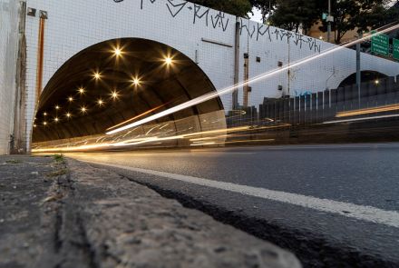 Vista em ângulo baixo da entrada do túnel à noite. A entrada é um arco revestido por azulejos brancos pichados. Dentro do túnel, luzes amarelas criam um efeito de estrela. Rastros de luz de veículos, em tons de branco e laranja, entram e saem do túnel, cruzando a imagem na horizontal.