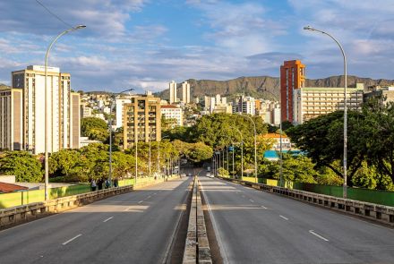 Vista a partir do centro de um viaduto com múltiplas pistas de asfalto. A via, vazia, se estende em perspectiva em direção a uma área urbana com prédios e muitas árvores. Ao fundo, uma cadeia de montanhas sob um céu azul. Postes de iluminação curvos ladeiam o viaduto.
