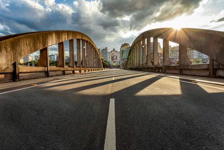 Vista em ângulo baixo a partir do meio da pista de asfalto do viaduto. Os dois grandes arcos de concreto da estrutura se erguem nas laterais, emoldurando a cena. O sol da tarde brilha intensamente à direita, projetando longas sombras dos pilares sobre o asfalto em direção ao observador.