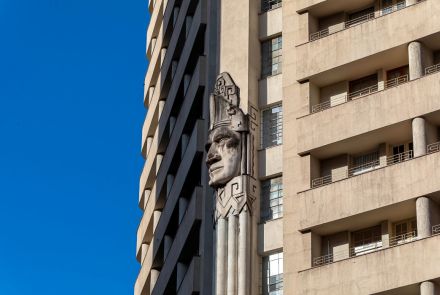 Um close na lateral do Edifício Acaiaca, mostrando um detalhe arquitetônico em relevo. No centro, uma escultura gigante de uma cabeça indígena esculpida em pedra ou concreto se destaca. A cabeça, com um cocar, está em uma parede estreita entre duas seções diferentes do prédio.
