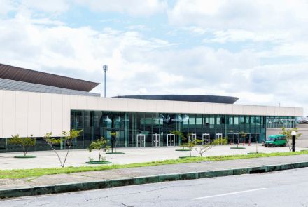 Fachada frontal de um edifício moderno, com paredes claras na parte superior e amplas janelas de vidro na parte inferior. Há várias portas de entrada enfileiradas sob a área envidraçada. Em frente ao prédio, existe um espaço pavimentado com pequenas árvores plantadas em canteiros circulares. À direita, um veículo verde está estacionado próximo à entrada. No primeiro plano, vê-se parte da via asfaltada e da calçada. Ao fundo, aparecem estruturas do telhado e uma torre metálica de iluminação