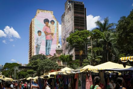 Imagem de uma feira ao ar livre com barracas de toldos listrados amarelo e branco. Pessoas circulam entre roupas expostas para a venda. Ao fundo, um prédio alto exibe um grande mural com a pintura de uma mulher negra carregando um bebê e duas crianças ao lado. Árvores cercam a cena.