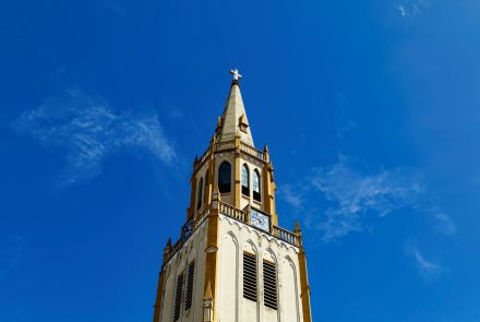 A torre pontiaguda da igreja sobe em destaque contra o céu limpo. Vemos janelas altas e estreitas, um relógio azul e a cruz no topo. A iluminação ressalta o tom amarelo claro da fachada.