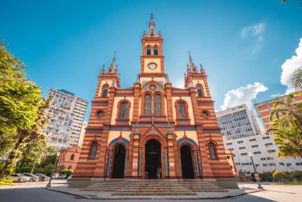 Fachada frontal e simétrica da Igreja de São José em Belo Horizonte. A igreja, de estilo histórico, é de cor vermelha e amarela, com três torres pontiagudas, sendo a central a mais alta e com um relógio. Uma escadaria leva à entrada principal. Ao lado, há prédios altos e árvores, sob um céu azul.
