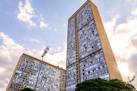 Dois edifícios altos de arquitetura modernista, com fachada envidraçada e laterais em concreto bege. O prédio da direita é mais alto e domina a cena, enquanto o da esquerda aparece parcialmente. Ao fundo, o céu está claro com algumas nuvens, e na parte inferior há árvores e um poste de iluminação urbana.