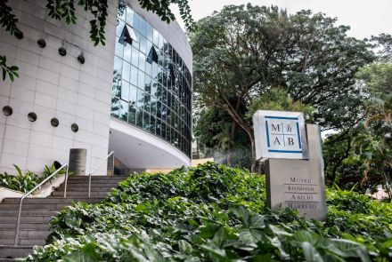 A entrada moderna do Museu Histórico Abílio Barreto. Em primeiro plano, há um canteiro com plantas verdes e a placa do museu, que exibe o nome e a sigla MHAB. O prédio curvo tem a fachada de concreto cinza e uma grande parede de vidro, que reflete as árvores. Uma escadaria de cimento leva à entrada do edifício.